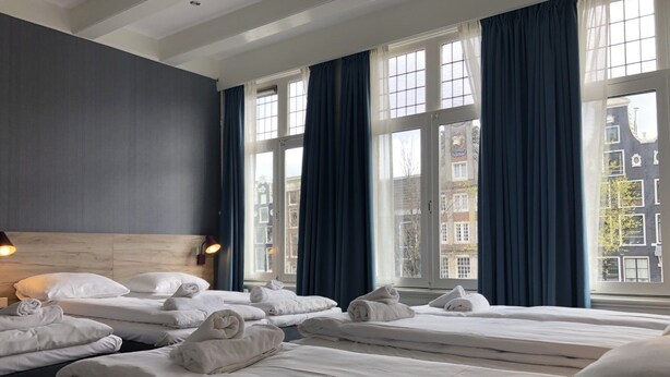 Quarto de hotel com três camas, cortinas escuras e vista para prédios através de janelas amplas.