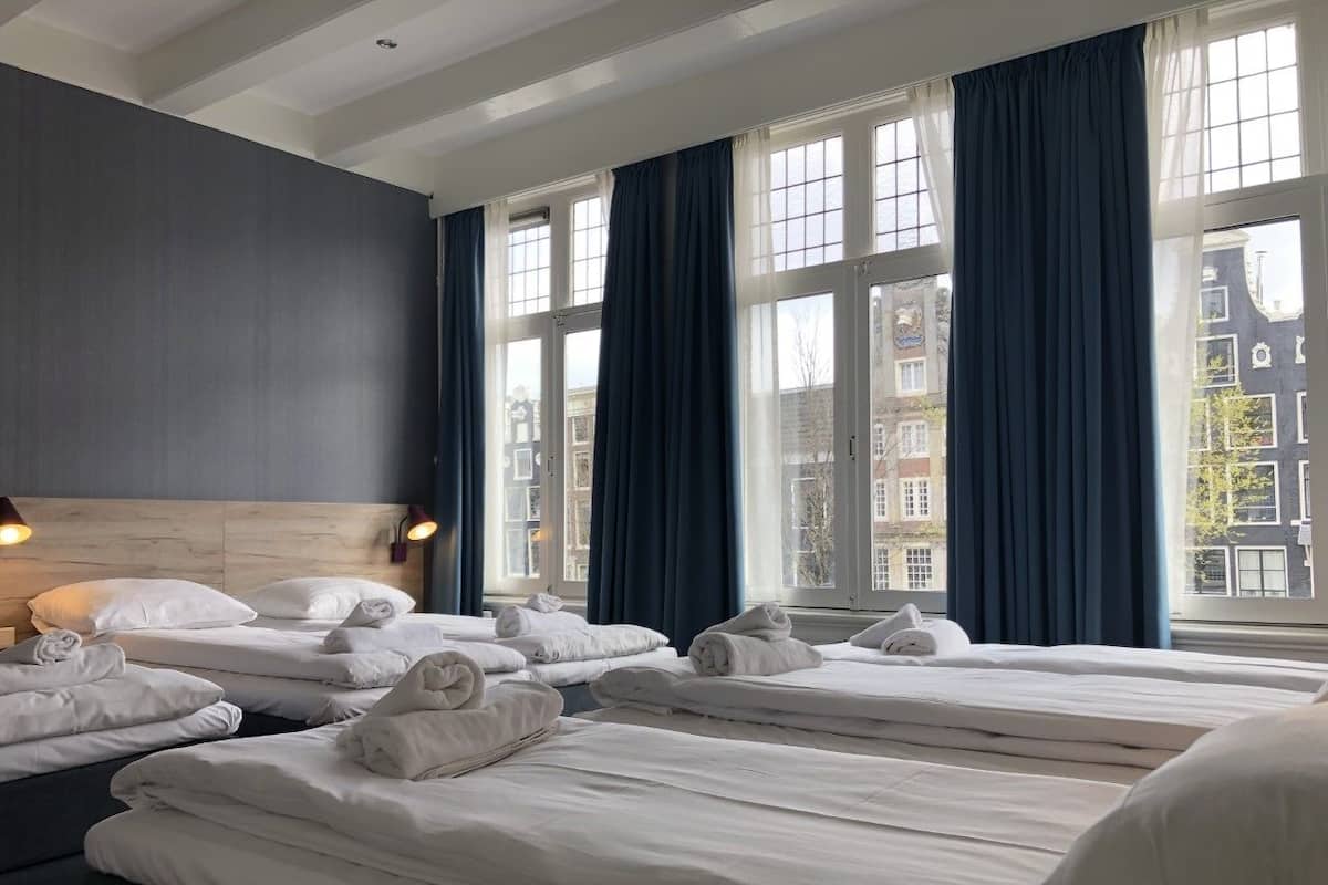 A hotel room with three beds, dark curtains, and a view of buildings through large windows.