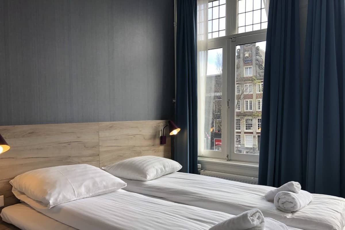 A hotel room with a large bed, two pillows, a headboard, a nightstand, a lamp, and a window with curtains.