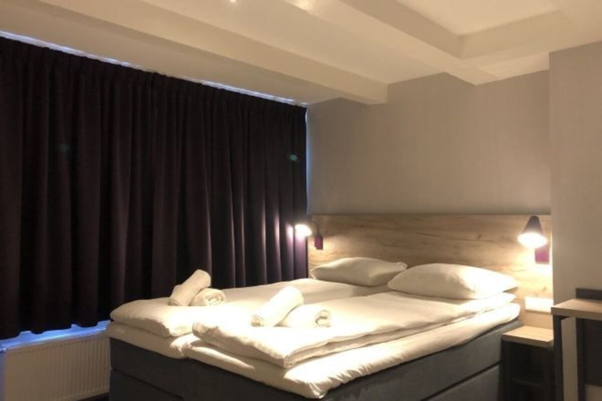 Habitación de hotel con cama, mesita de noche, lámpara y ventana con cortinas.