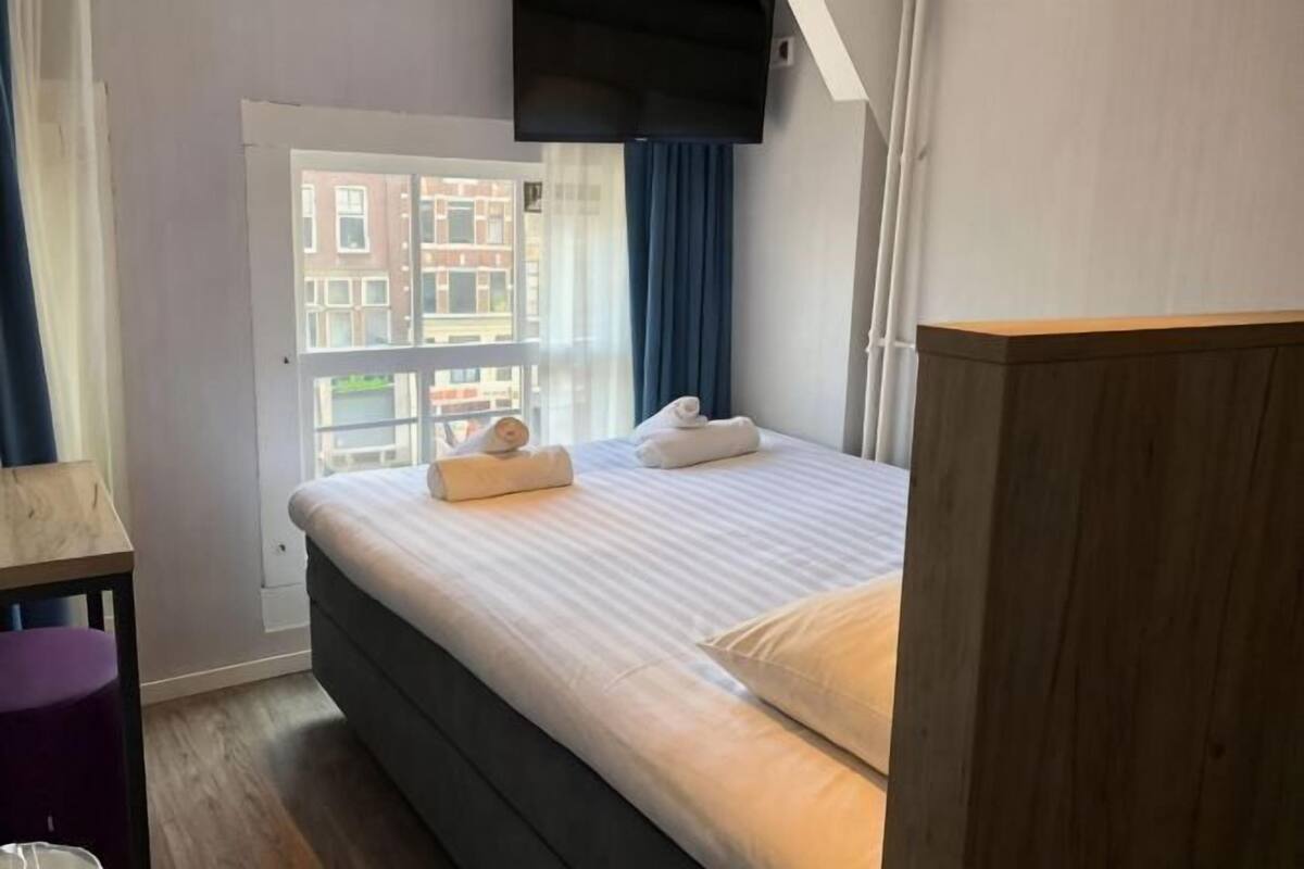 A bedroom with a bed, a wooden headboard, a window with curtains, and a view of buildings outside.