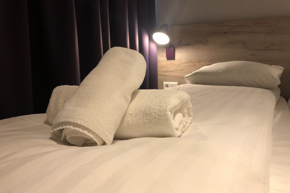 A neatly made bed with white linens and rolled towels, a wooden headboard, and a wall-mounted light fixture.