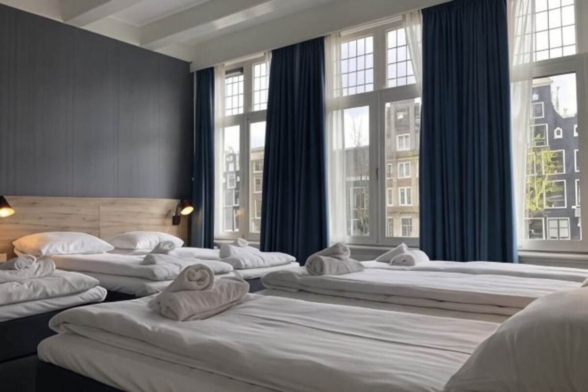 Habitación de hotel con varias camas, ventanas amplias y vistas a edificios.