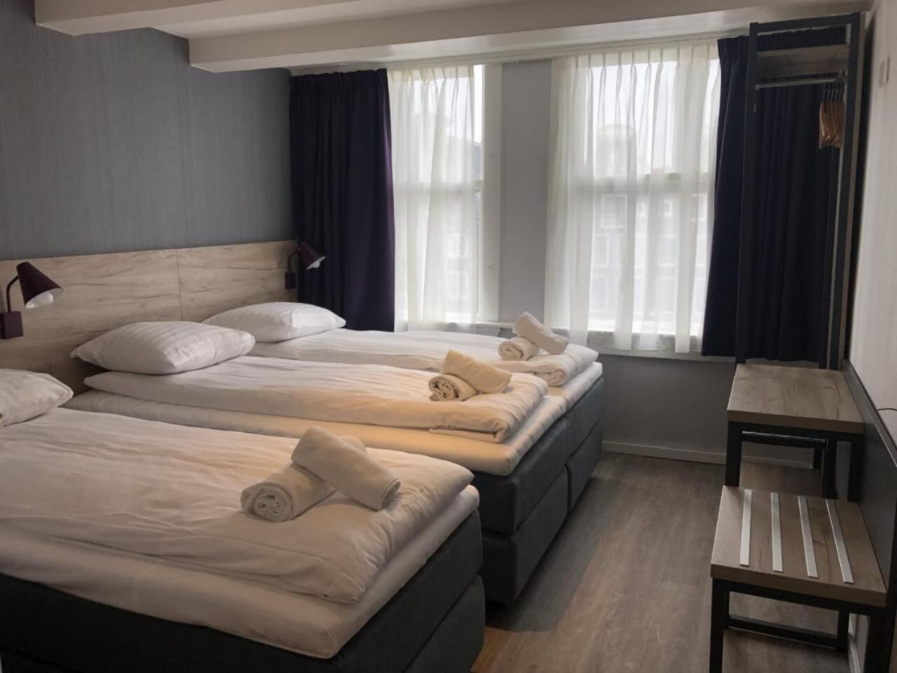 Habitación de hotel con tres camas, una mesita pequeña y un ventanal con cortinas.