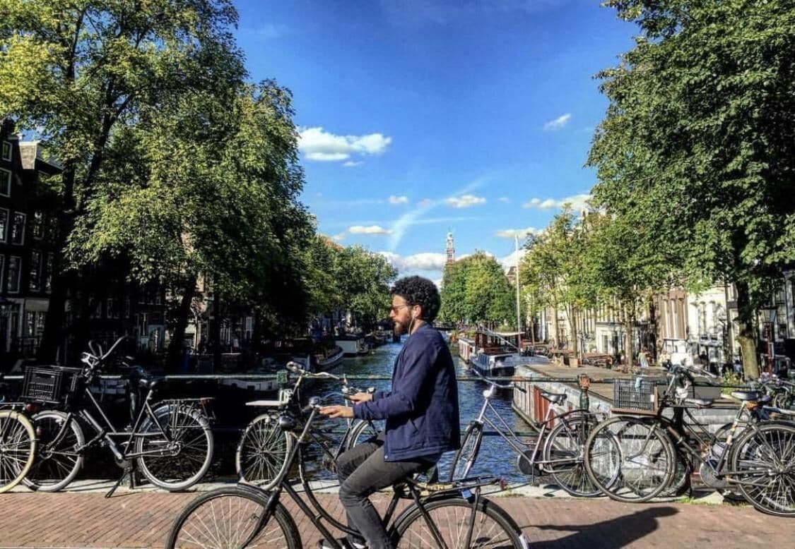 Eine Person fährt mit dem Fahrrad entlang eines kanalähnlichen Wasserwegs, gesäumt von Bäumen, mit Gebäuden an beiden Seiten.