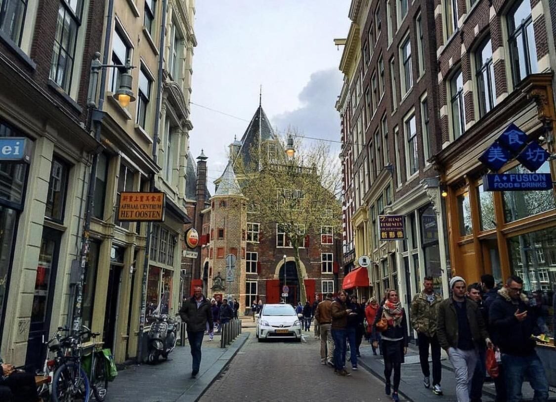 Eine schmale Straße mit Fußgängern, Fahrrädern und einem weißen Auto. Gebäude an beiden Seiten zeigen Werbeschilder, im Hintergrund dominiert ein markanter Kirchturm.