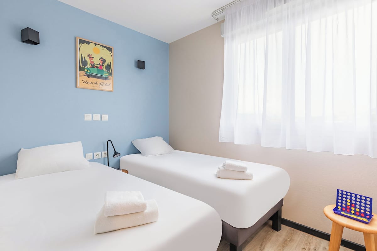 A hotel room with two beds, a painting on the wall, a small table with a board game, and a window with sheer curtains.