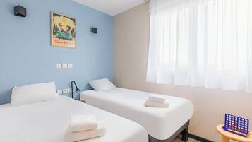 A hotel room with two beds, a painting on the wall, a small table with a board game, and a window with sheer curtains.