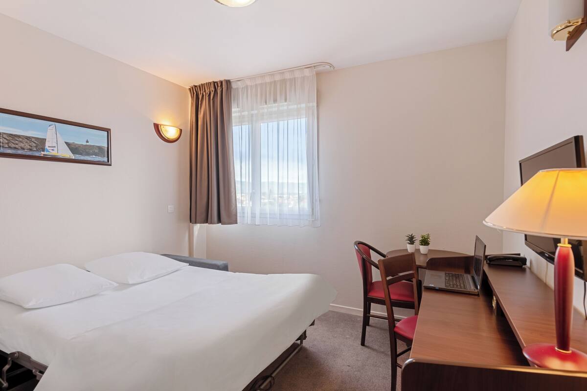 A hotel room with a bed, a desk with a laptop, a chair, a lamp, a TV, and a window with curtains.