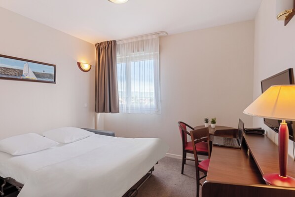 Una habitación de hotel con una cama, un escritorio con una computadora portátil, una silla, una lámpara, un televisor y una ventana con cortinas.