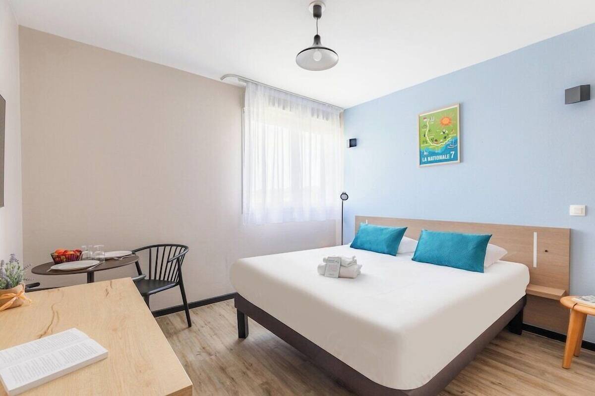 A hotel room with a bed, a desk, a chair, a small table, a plant, and a window with curtains.