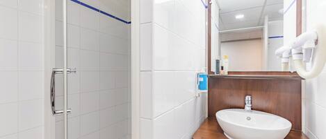 Un baño moderno con paredes de azulejos blancos, un tocador de madera, un lavabo blanco y un box de ducha de vidrio.