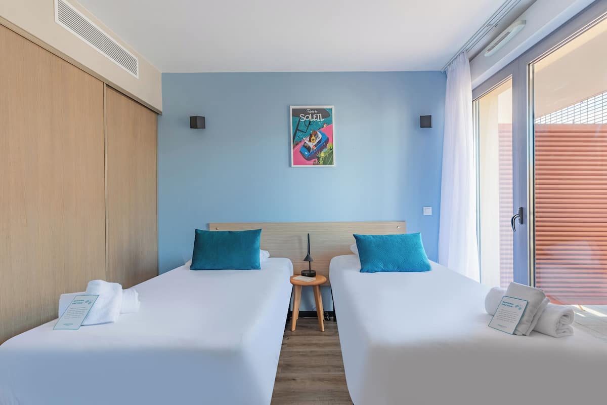 A hotel room with two beds, a small table, a poster on the wall, and a window with blinds.