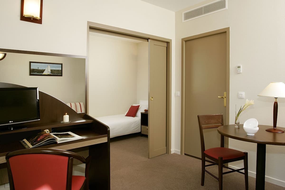 A hotel room with a bed, a desk with a TV, a chair, a table with a lamp, and a door leading to another room.