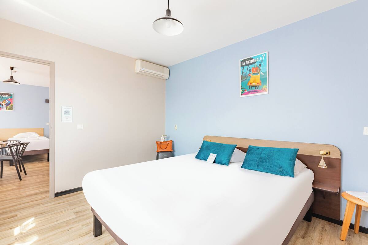 A hotel room with a large bed, a desk, and a chair. There is a poster on the wall and an air conditioning unit.