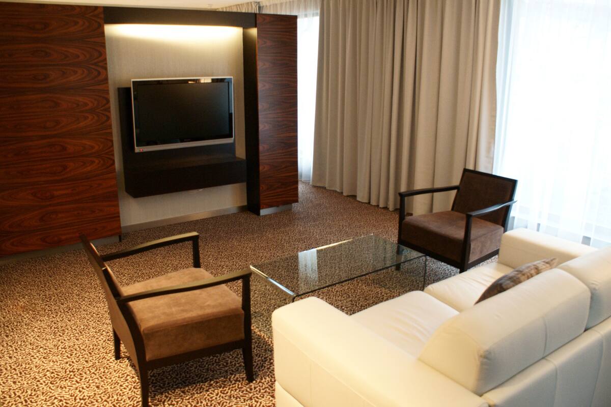 A modern living room with a flat-screen TV mounted on a wooden panel, a glass coffee table, a white sofa, and two brown armchairs.