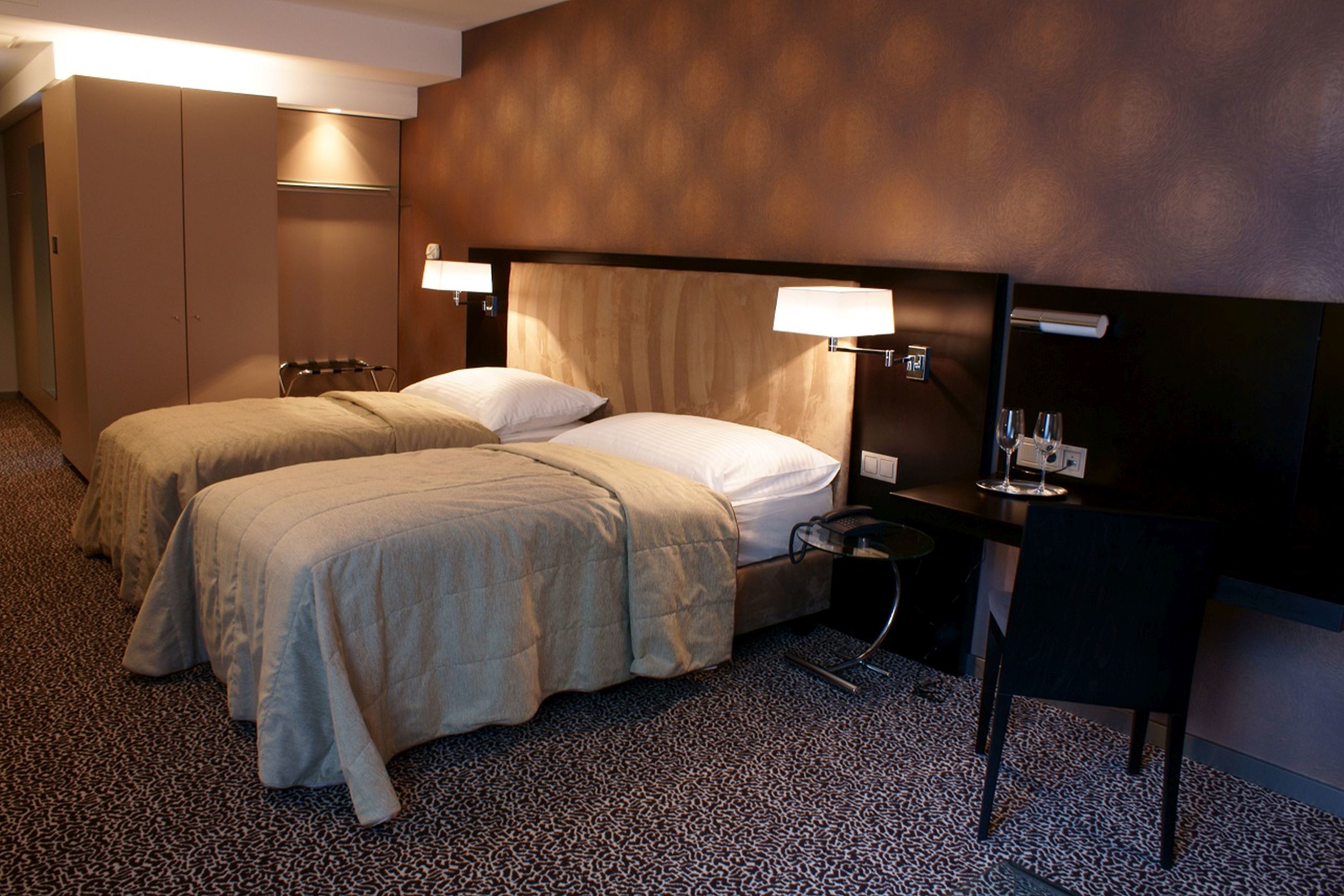 Habitación de hotel con dos camas, un escritorio y una silla. Hay lámparas en el escritorio y en la pared, y un armario en la esquina.