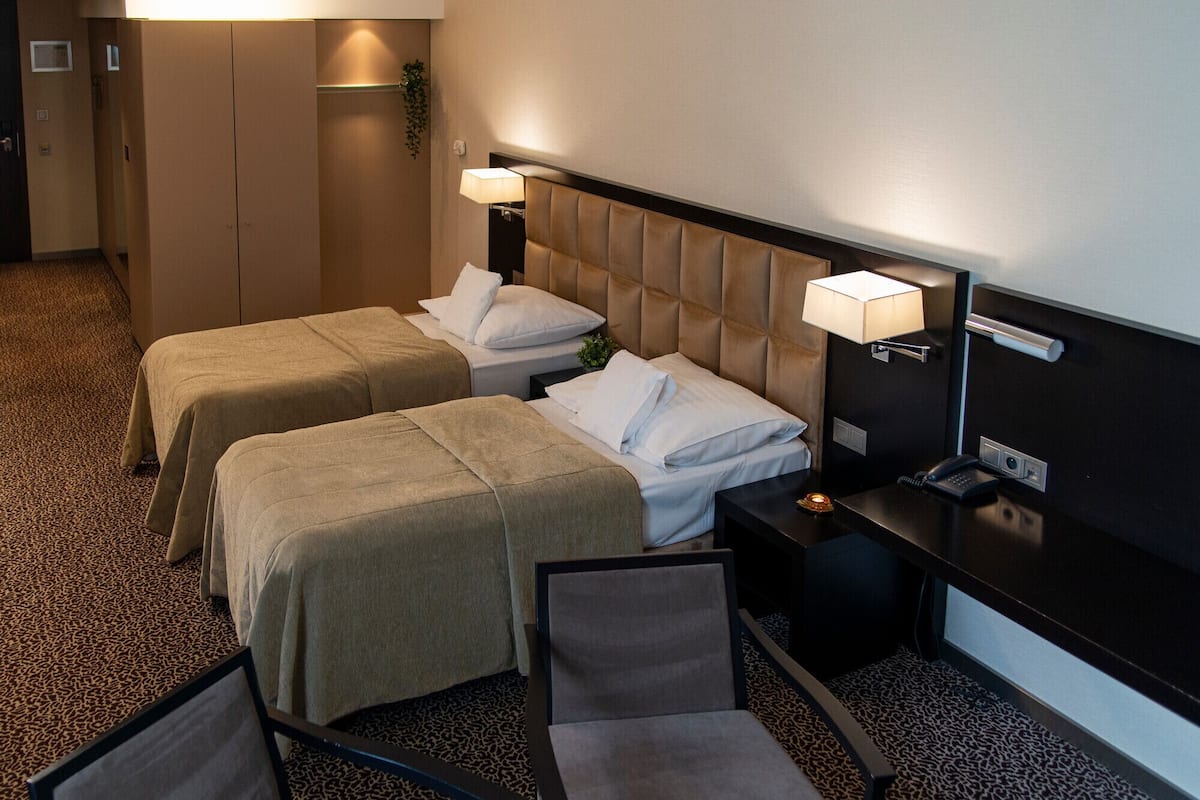 A hotel room with two beds, a desk, a chair, and a nightstand with a lamp.