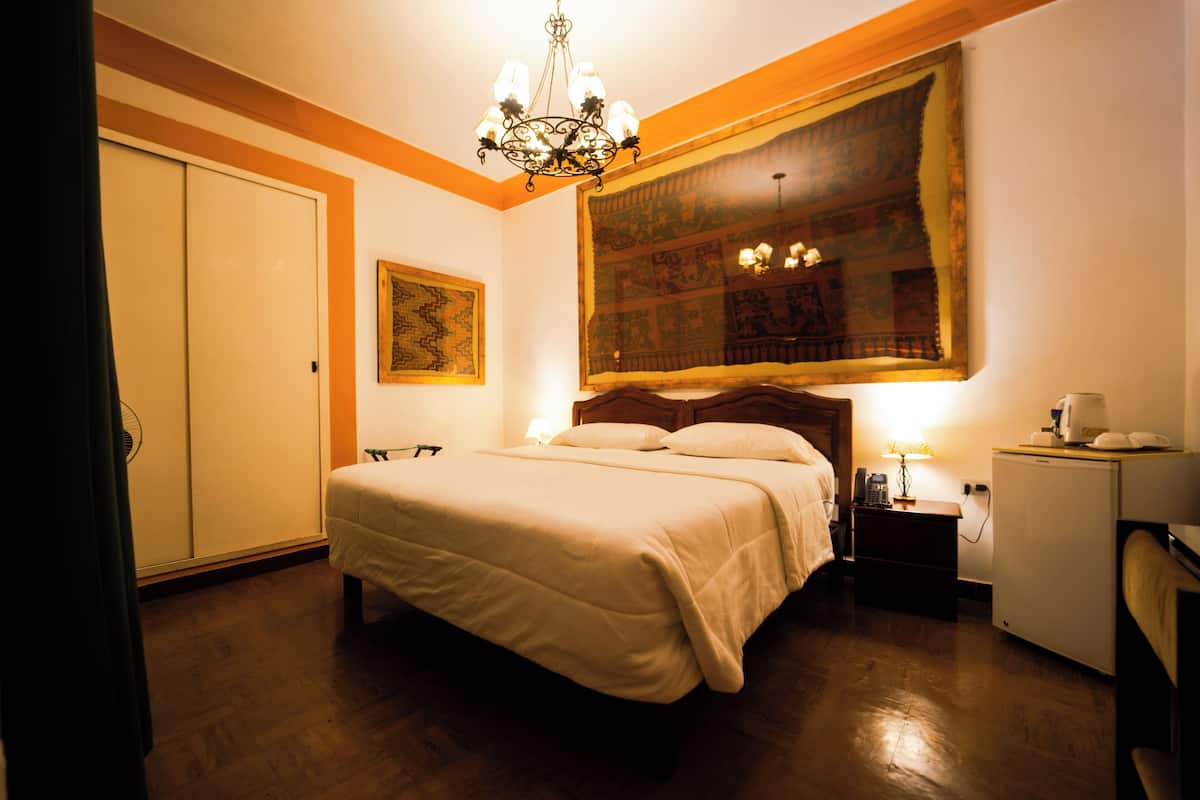A hotel room with a large bed, a nightstand with a lamp, a small refrigerator, and a large tapestry on the wall.