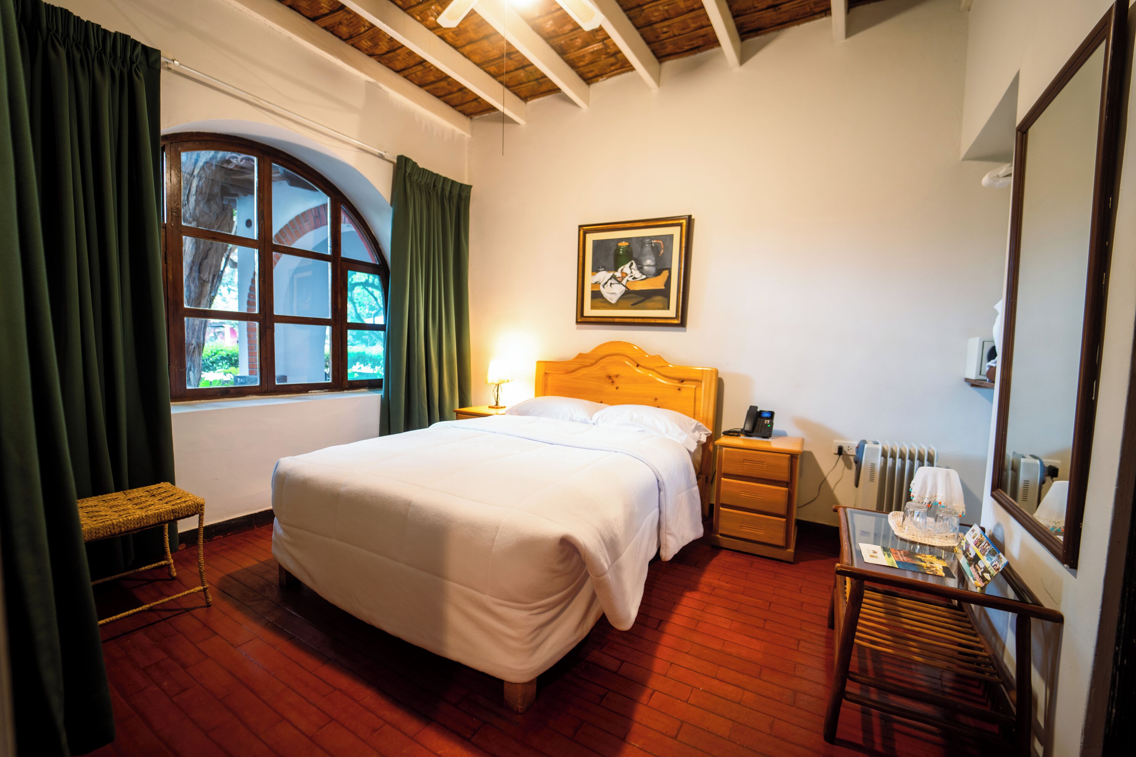 Un dormitorio con una cama grande, cabecera de madera, mesita de noche, una pequeña mesa con lámpara, ventana con cortinas e imagen enmarcada en la pared.
