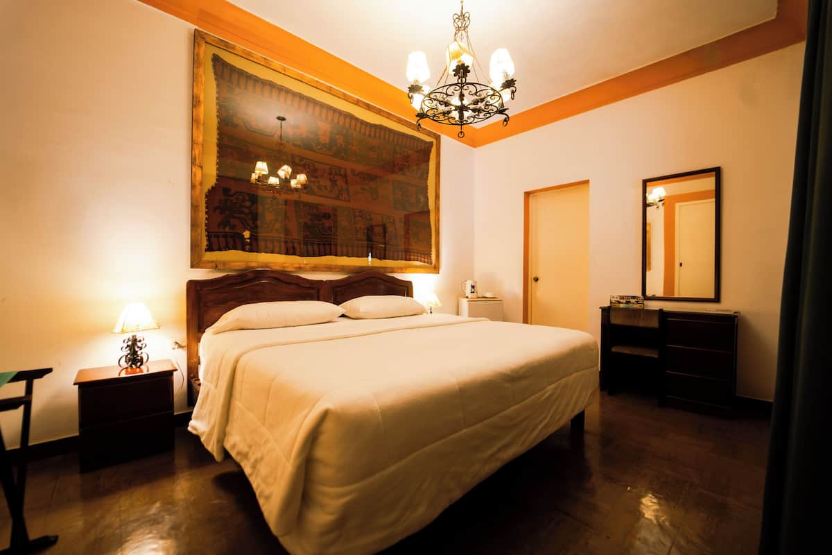 A hotel room with a large bed, a wooden headboard, a nightstand with a lamp, a dresser, and a large mirror.