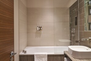 Un baño moderno con bañera, lavamanos y zona de ducha. Hay artículos de aseo en la pared y una toalla colgada al lado de la bañera.