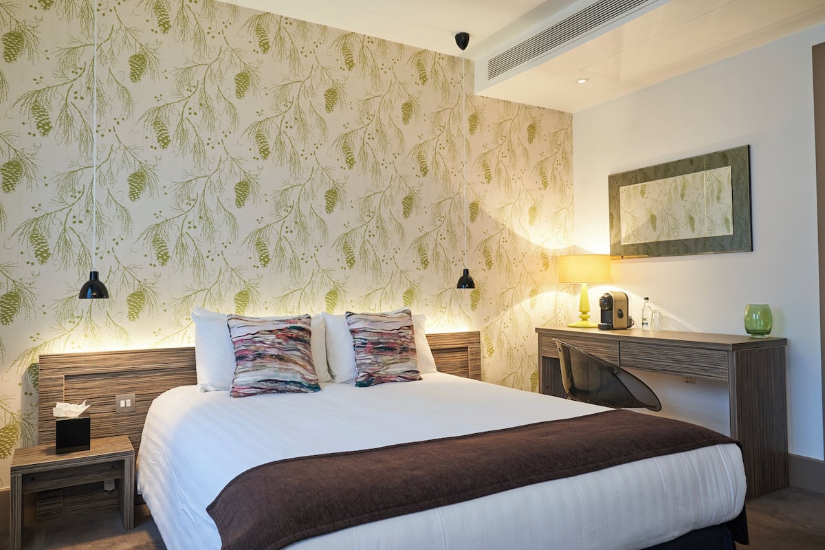 Quarto de hotel com uma cama grande, duas mesas de cabeceira, um criado-mudo, uma escrivaninha com luminária e um espelho fixado na parede.