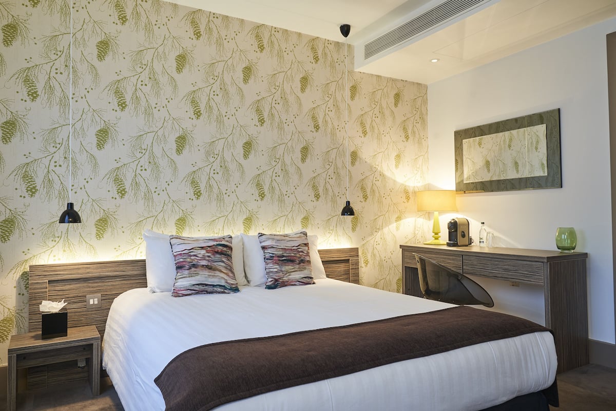 Quarto de hotel com uma cama grande, uma mesa de cabeceira, uma escrivaninha com abajur e um espelho fixado na parede.