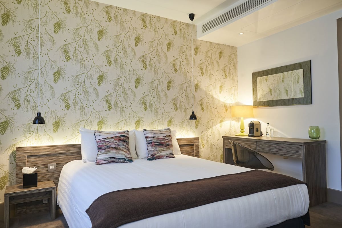 Quarto de hotel moderno com uma cama grande, duas mesas de cabeceira, um criado-mudo, uma escrivaninha com abajur e um espelho fixado na parede.