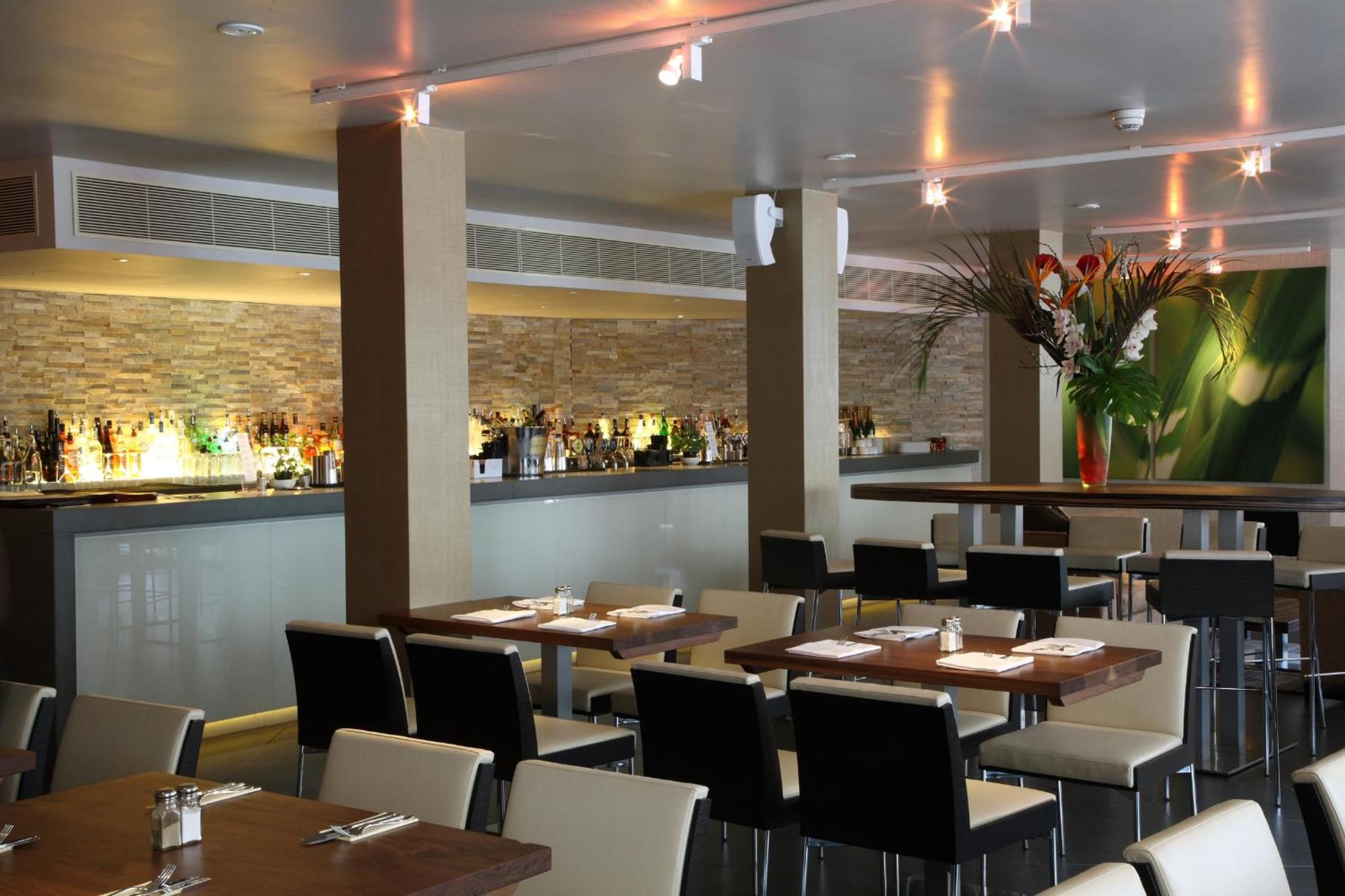 A restaurant with multiple tables and chairs, a bar area with bottles, and a decorative plant.