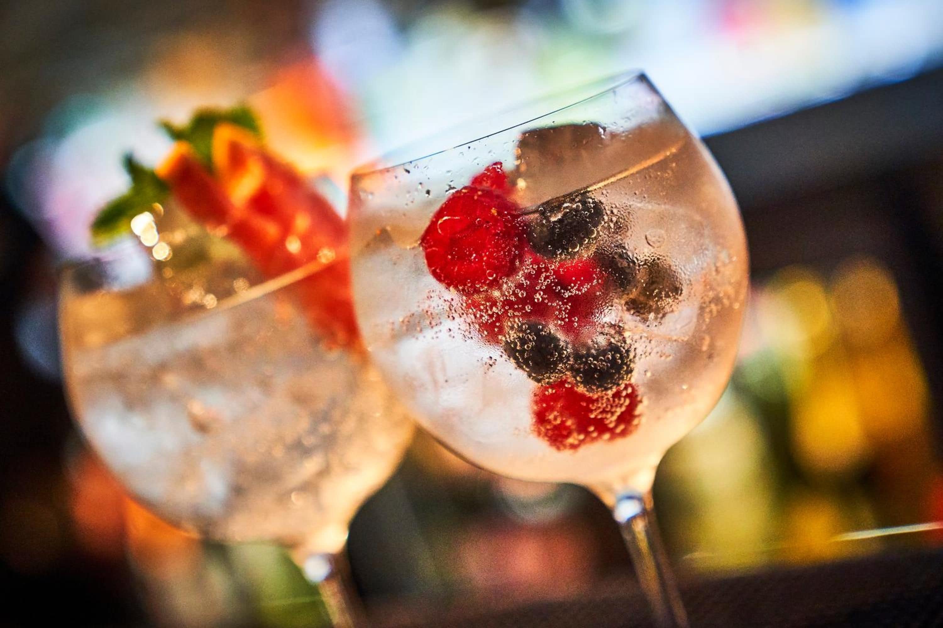 Two glasses of a clear beverage with ice cubes and garnishes, likely a cocktail, with a blurred background suggesting a bar or restaurant setting.