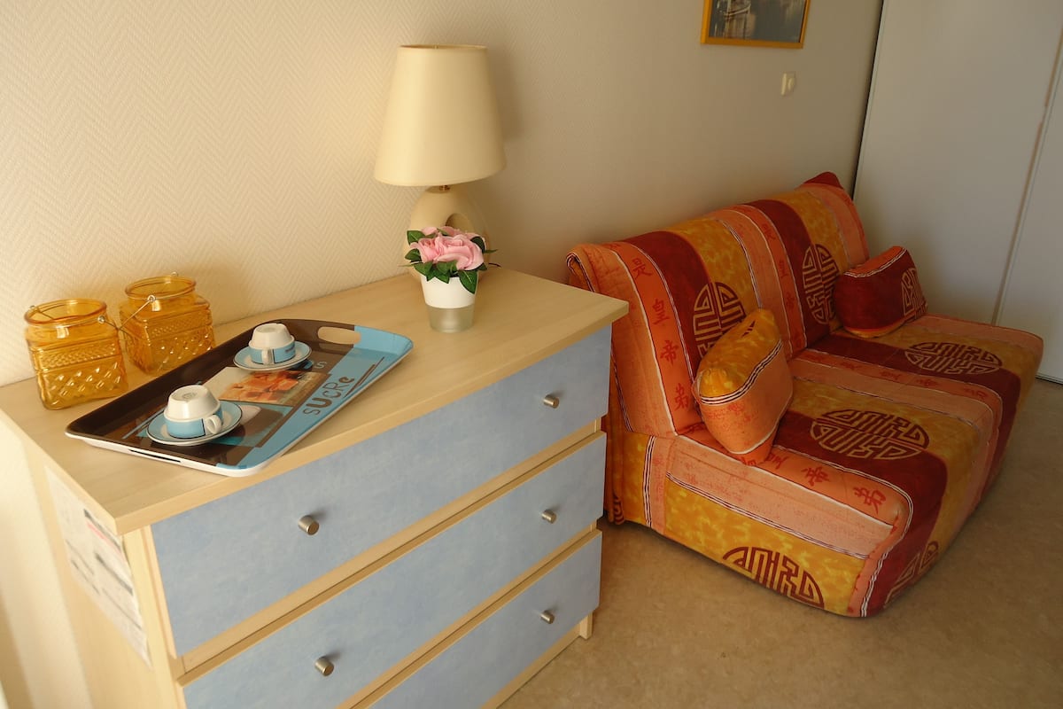 Quarto com cômoda de madeira, bandeja com xícaras de chá, luminária e um sofá colorido.