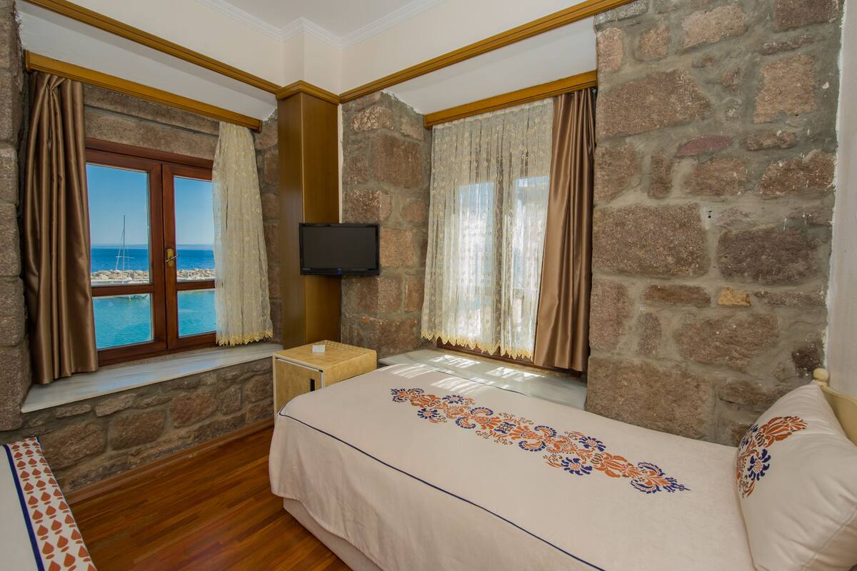A bedroom with a stone wall, a bed with patterned bedding, a window with curtains, and a TV mounted on the wall.
