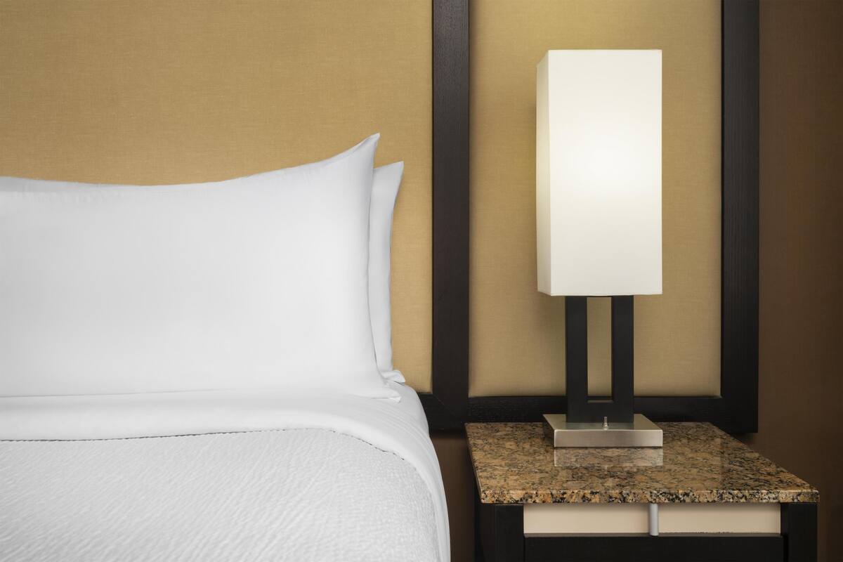 A neatly made bed with white linens and a modern bedside table with a lamp.