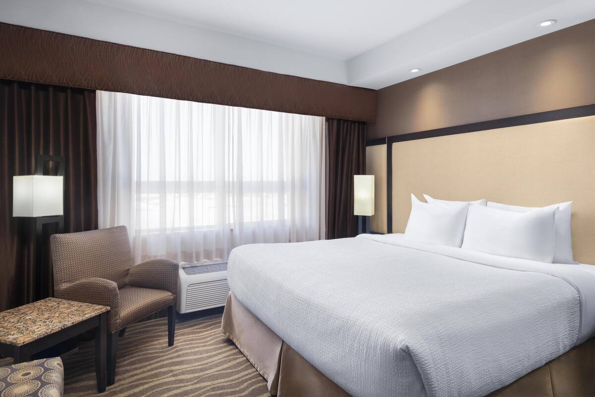 A hotel room with a large bed, a chair, a small table, and a large window with curtains.
