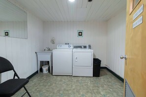 A laundry room with a washing machine, dryer, and a sink. There is a chair and a trash bin.