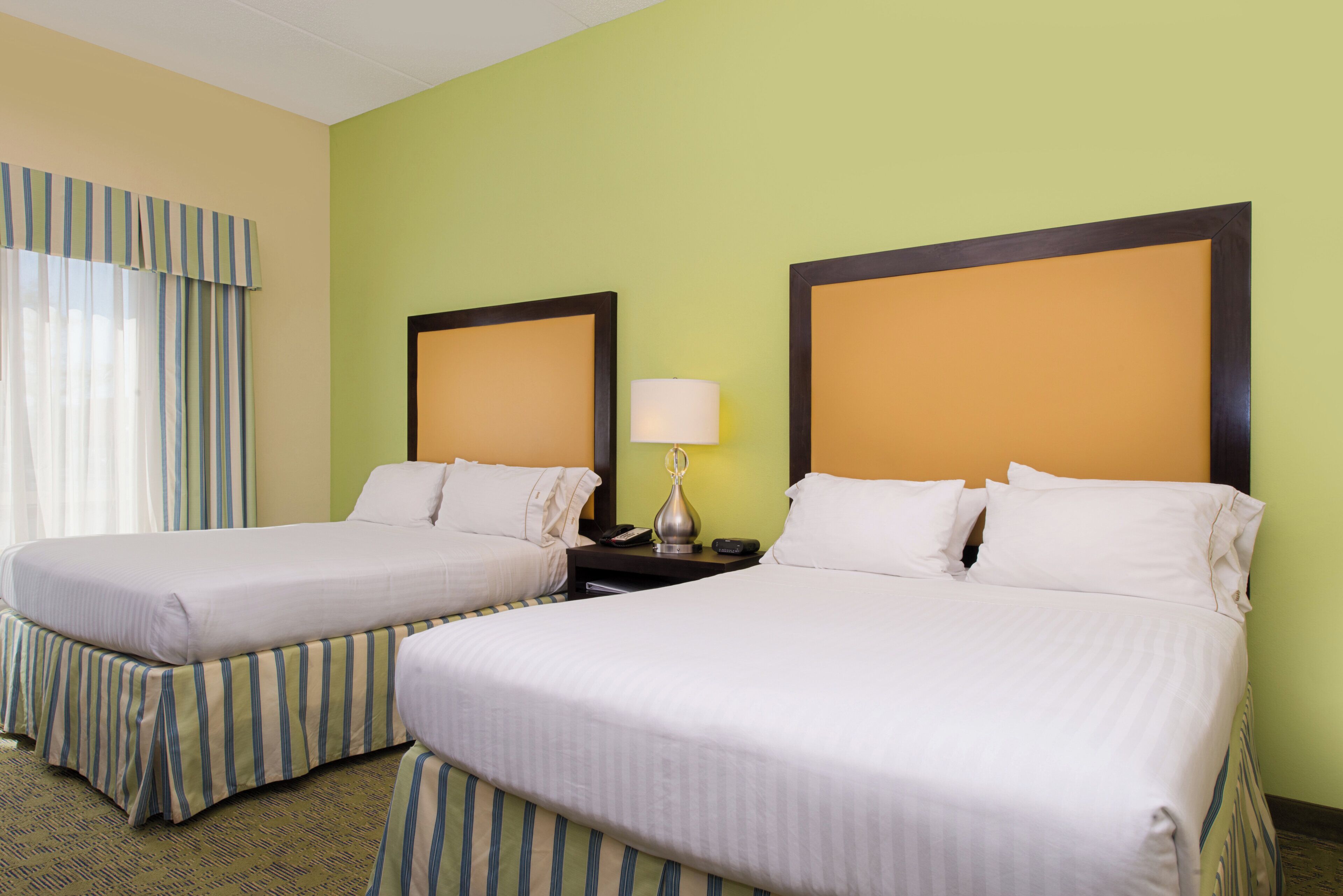 A hotel room with two beds, a nightstand with a lamp, and a window with curtains.