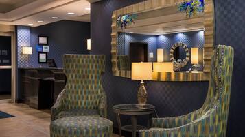 Un hall de hotel con una recepción, dos sillones tapizados, una mesa redonda con una lámpara y un espejo grande en la pared.