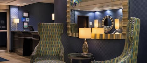 Un hall d’hôtel comprenant un comptoir d’accueil, deux fauteuils rembourrés, une table ronde avec une lampe et un grand miroir au mur.