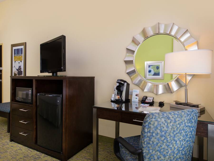Habitación de hotel con escritorio, televisión, microondas, refrigerador, silla, lámpara y espejo.