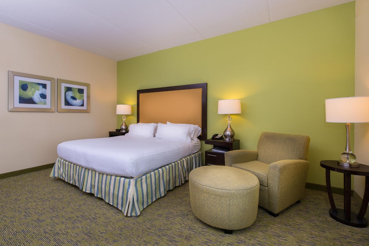 A hotel room with a bed, a chair, an ottoman, two bedside tables, and two wall-mounted lamps.