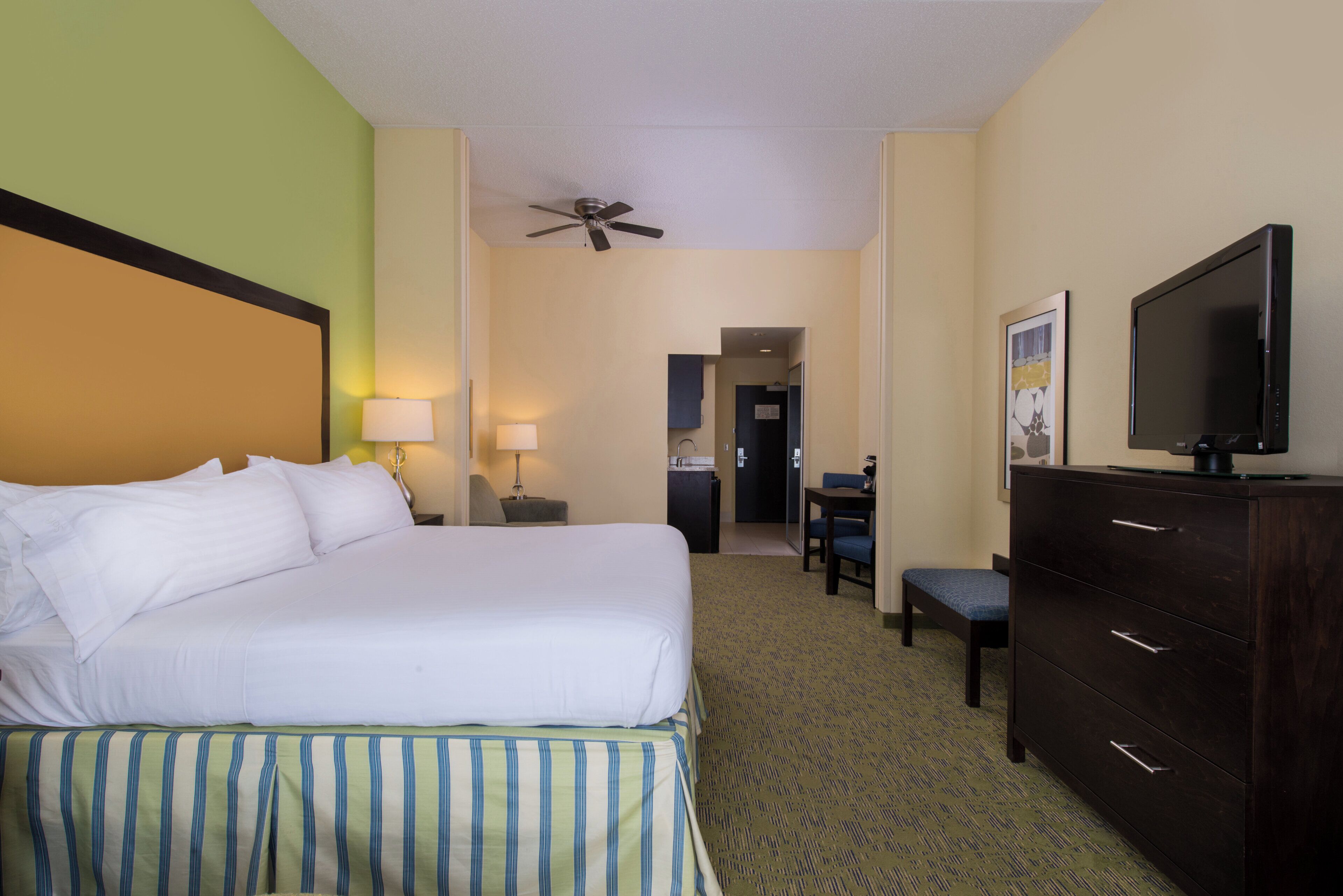 A hotel room with a bed, a desk, a chair, a dresser, a television, and a ceiling fan.