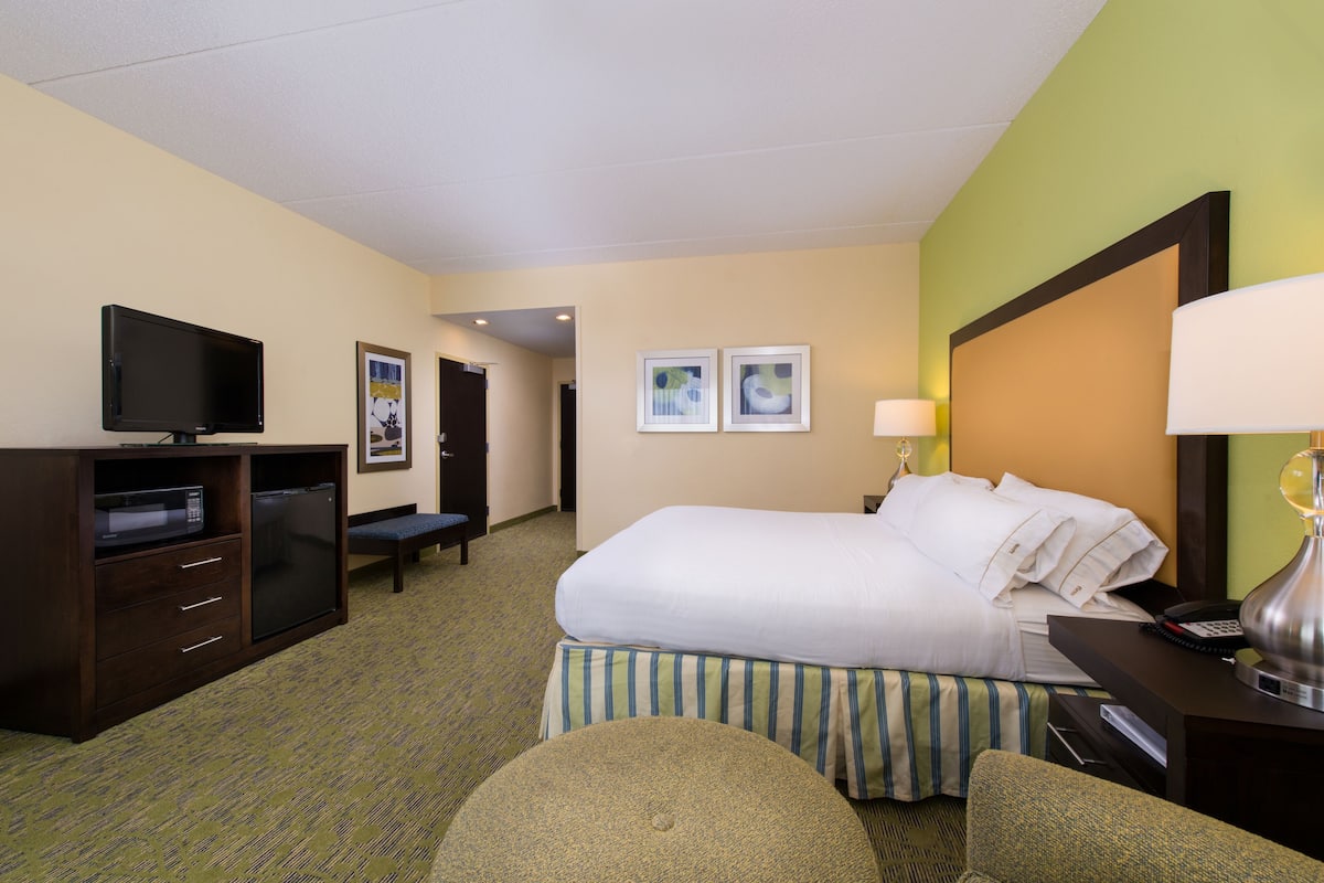 A hotel room with a bed, a desk with a lamp, a television, a minibar, and two framed pictures on the wall.