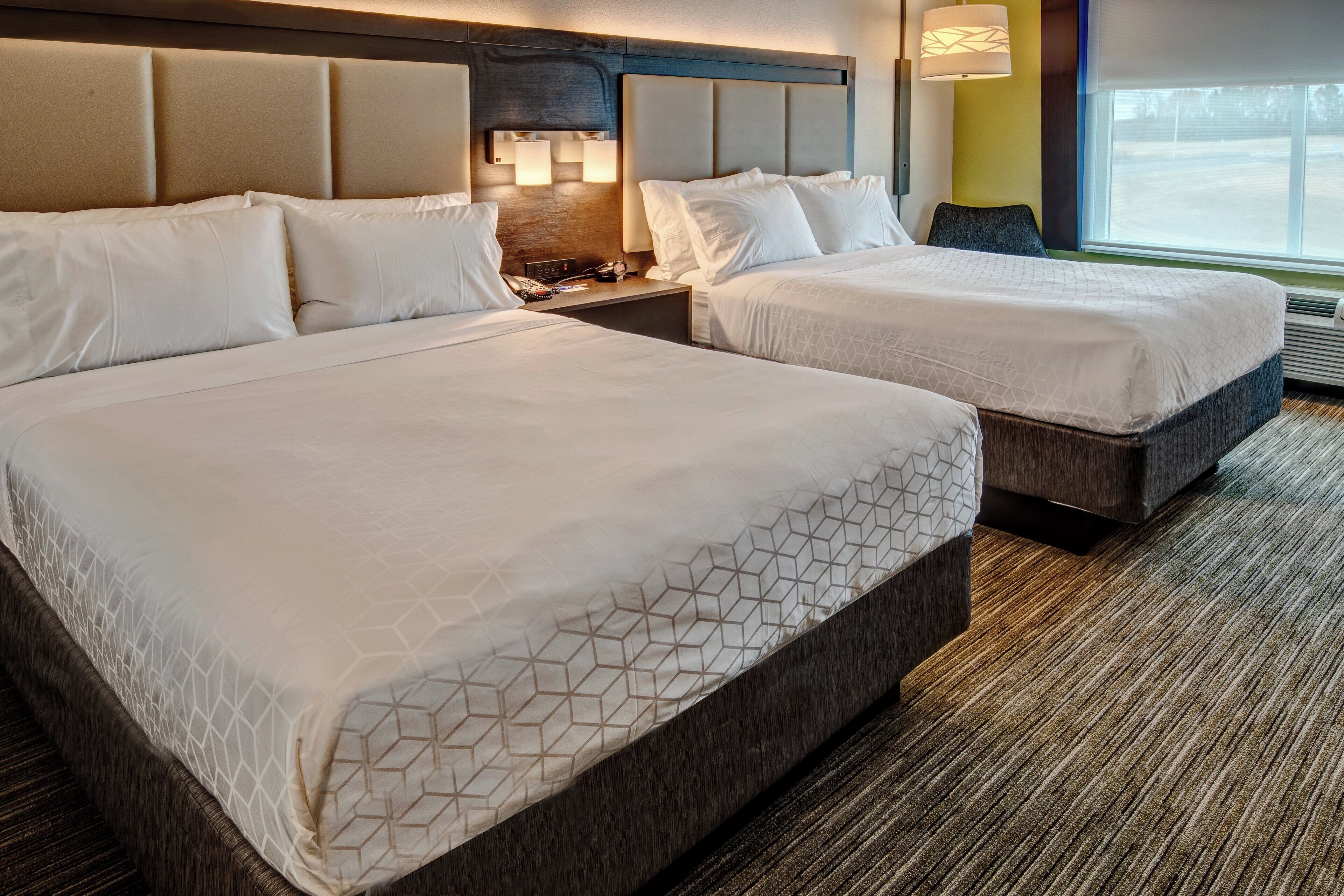 A hotel room with two beds, a headboard with built-in lighting, a bedside table with a lamp, and a view of the outdoors through a window.