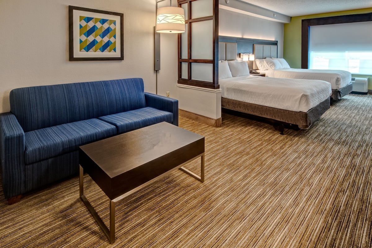 Une chambre d’hôtel avec un canapé bleu, une table basse en bois, deux lits et une grande fenêtre.