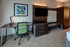 A hotel room with a desk, chair, TV, wardrobe, and a framed artwork on the wall.