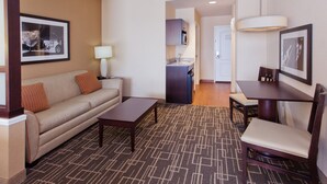 A hotel room with a sofa, a coffee table, a dining table with chairs, a kitchen area, and a framed picture on the wall.