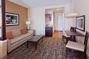 A hotel room with a sofa, a coffee table, a dining table with chairs, a kitchen area, and a framed picture on the wall.