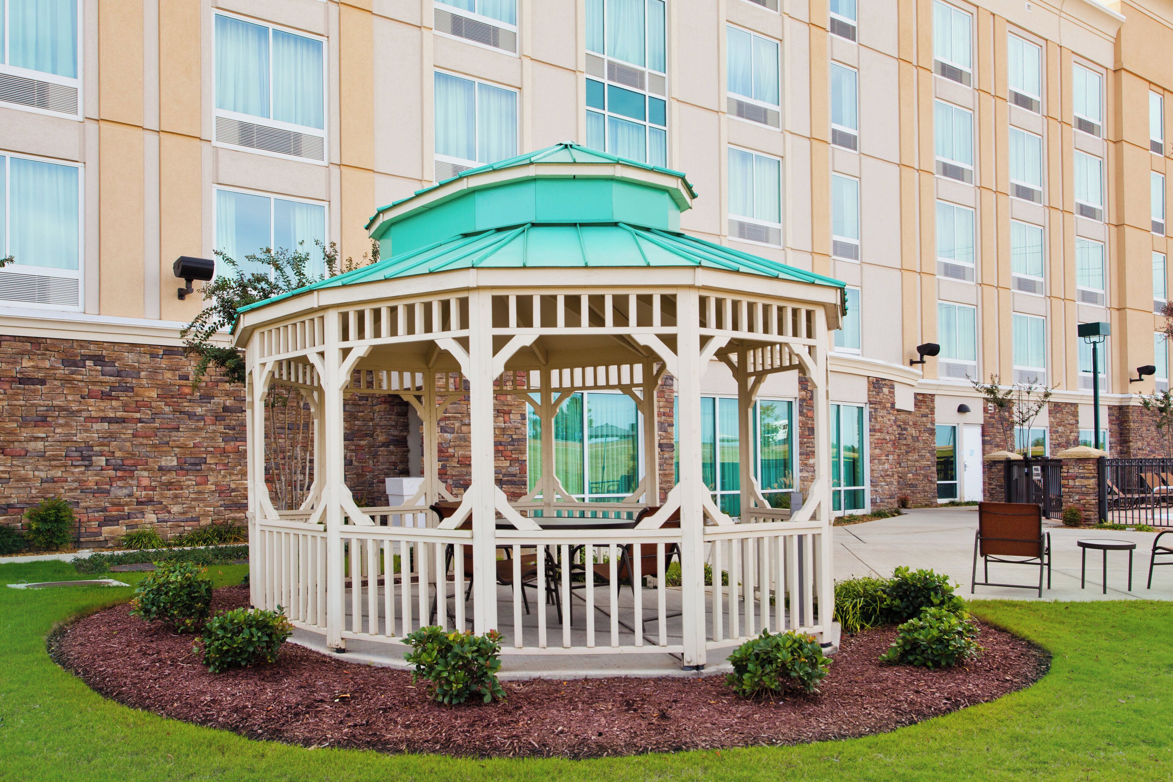 A gazebo with a green roof and white walls, surrounded by a landscaped garden with small bushes.