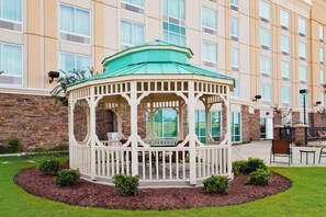 A gazebo with a green roof and white walls, surrounded by a landscaped garden with small bushes.